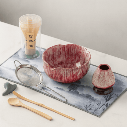 Red ceramic bowl with matching small pitcher on a decorative mat with tea-making tools.