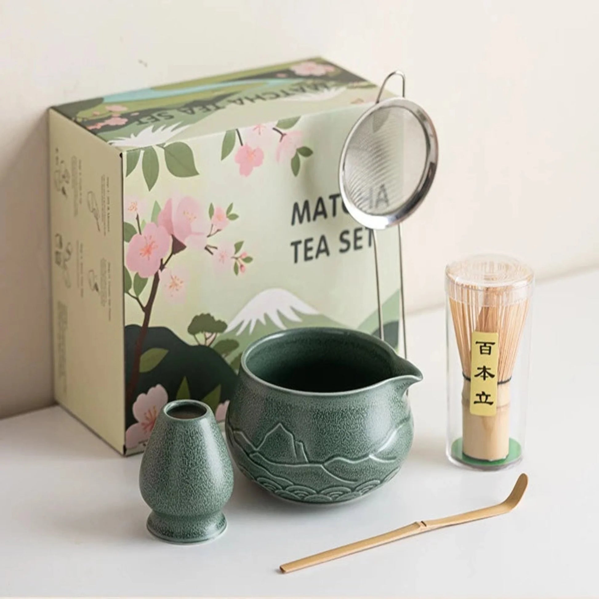 Matcha tea set with green teapot, whisk, and scoop on a white surface with a decorative box in the background.