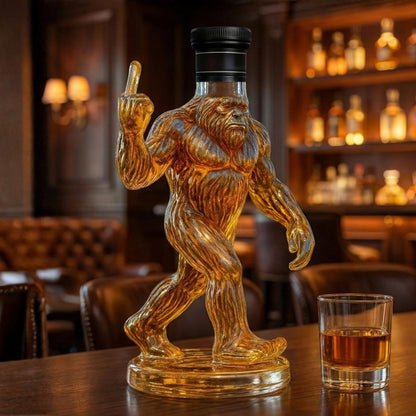 A handcrafted glass Bigfoot Sasquatch whiskey decanter filled with bourbon, giving the middle finger on a luxury bar top next to a glass of whiskey.